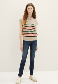 Haut en tricot beige avec un col zippé, comportant des rayures horizontales en rouge, vert et marine. Associé à un jean bleu foncé et des mocassins compensés crème.