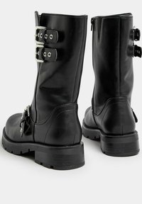 Las botas de tobillo de cuero negro cuentan con dos hebillas plateadas, un cierre de cremallera y una suela de goma texturizada con un tacón grueso para mayor estabilidad.