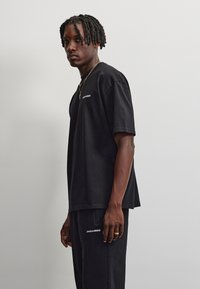 Ung mand med korte dreadlocks iført sort Pegador t-shirt og bukser, stående sidelæns mod en ensfarvet lys baggrund.