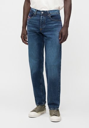 Uomo che indossa jeans blu a gamba dritta, camicia bianca infilata e sneakers verde oliva in piedi davanti a uno sfondo uniforme.
