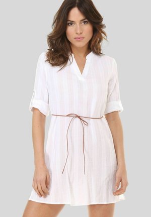 Mujer con cabello castaño ondulado, lleva un vestido blanco hasta la rodilla con mangas remangadas y un delgado cinturón marrón atado a la cintura.