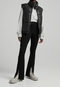 Musta vuorellinen liivi harmaan collegepaidan päällä, yhdistettynä mustiin ribbihousuihin, joissa on sivureiät, ja beigekenkäisiin nauhaliivikengiin.