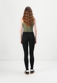 Schwarze enge Jeans mit hoher Taille, strukturiertes olivgrünes Tanktop und Gesäßtaschen. Die Schuhe haben schwarze und weiße Akzente mit einer Sohle.