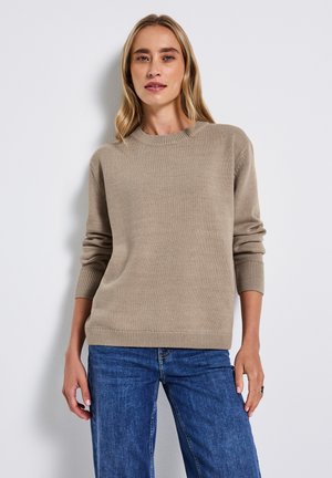 Beige striktrøje med rund hals, ribstrikkede manchetter og kant, parret med blå jeans. Stoffet ser glat og medium vægtigt ud.