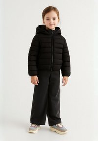 Jachetă puffer neagră cu glugă, având cusături orizontale și fermoar în față. Asortată cu pantaloni negri cu talie înaltă și adidași.