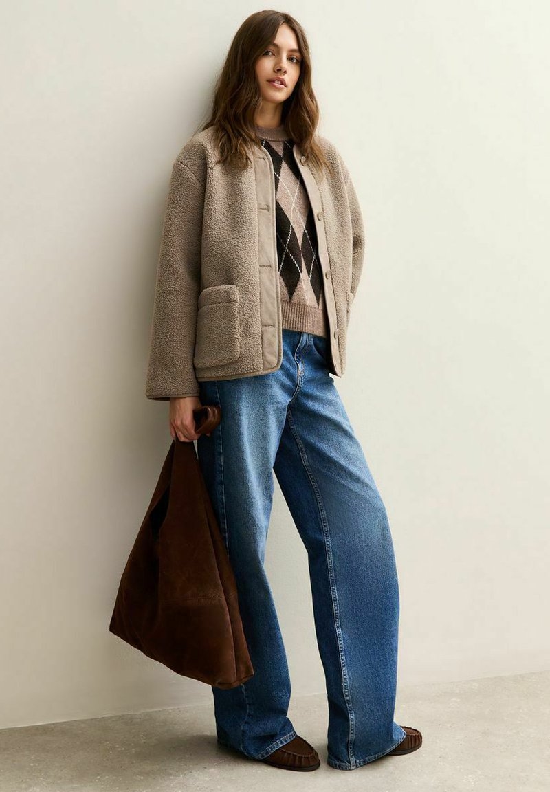 Beige jas met fleecevoering en grote knopen, gecombineerd met een trui met ruitpatroon en blauwe wijde jeans. Brown suède tas en mocassins.