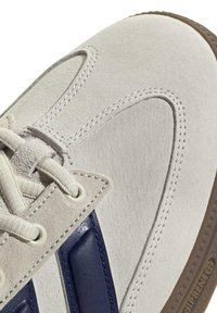 Gros plan sur l’avant d’une basket en daim beige avec lacets blancs, coutures visibles, détail en cuir bleu marine et bord de semelle en caoutchouc marron texturé.