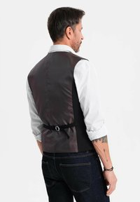 Mann in einem hochgekrempelten gestreiften Hemd, schwarzem Satinrückenweste mit verstellbarem Träger und dunklen Jeans, von hinten vor einem weißen Hintergrund dargestellt.