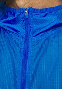 Niebieska lekka kurtka z niebieskim zamkiem błyskawicznym na środku, częściowo zapięta, odsłaniająca szyję i odcień skóry osoby powyżej kołnierza.