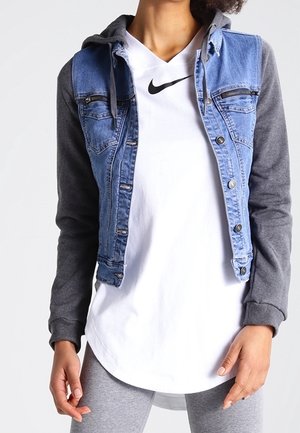 Person trägt ein weißes Nike-Shirt, graue Hoodie-Ärmel, blaue Jeansweste mit Knöpfen und graue Leggings, steht mit entspannten Händen.