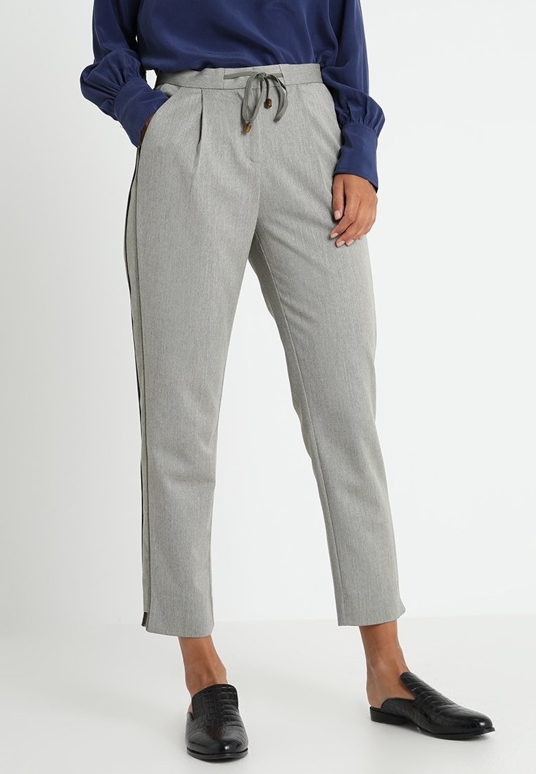 Femme portant un pantalon gris clair fuselé avec cordon de serrage et bande latérale, blouse bleu marine à manches longues et chaussures noires sans lacets.