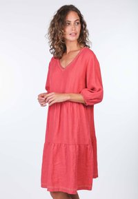 Korallenfarbenes Leinenkleid mit V-Ausschnitt, Dreiviertelärmeln, gestuftem Rüschensaum und Spitzenbesatz. Glatte Textur, entspannte Silhouette.