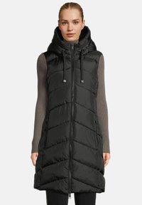 Woman wearing a black quilted sleeveless hooded vest over a long-sleeve gray ribbed shirt, standing against a plain background.