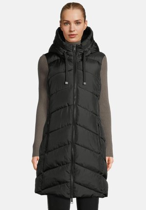Vrouw in een zwart gewatteerd mouwloos vest met capuchon over een grijze geribbelde longsleeve, staand tegen een plain achtergrond.
