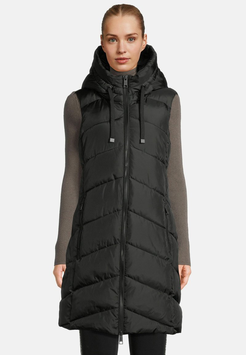 Woman wearing a black quilted sleeveless hooded vest over a long-sleeve gray ribbed shirt, standing against a plain background.
