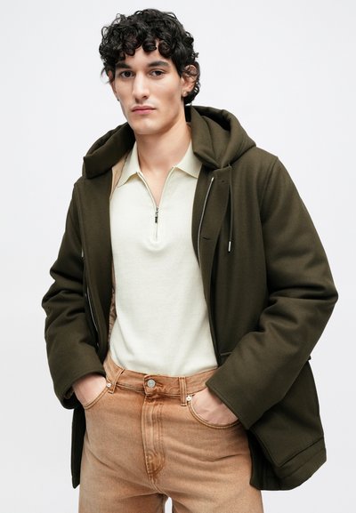 Oliivinvihreä hupullinen takki kreemivalkoisen vetoketjullisen polopaidan päällä, yhdistetty vaaleanruskeisiin farkkuihin. Sileät kankaat ja hillityt metalliosat.