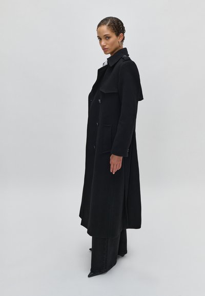 Juodas ilgas paltas su aukštu apykakle, pagamintas iš tekstūrinės medžiagos, su dviem priekinėmis kišenėmis ir sagų detalėmis, uždėtas ant tamsių džinsų.