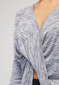 Šedá a černá vzorovaná blůza s výstřihem do V a stočeným předním detailem, vyrobená z měkké, texturované látky. Vhodná pro příležitostné nošení.