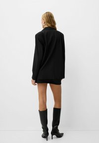 Blazer noir oversize avec un col structuré, associé à une mini jupe noire et des bottes stilettos en cuir jusqu'aux genoux. Texture lisse, design minimal.