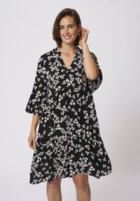 Robe noire à fleurs avec un décolleté en V, des manches évasées, et un design à étages. Présente de petites fleurs blanches et roses sur un tissu texturé.