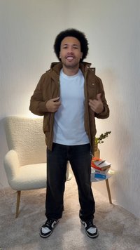 Brązowa kurtka na suwak na białej koszulce, czarne spodnie i czarno-białe trampki. W tle jasny fotel i książki.