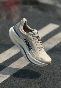 Λευκό αθλητικό παπούτσι Hoka με μπλε και πορτοκαλί λεπτομέρειες, αιωρείται πάνω από πεζοδρόμιο με άσφαλτο, με μικρά θραύσματα κάτω από την σόλα.