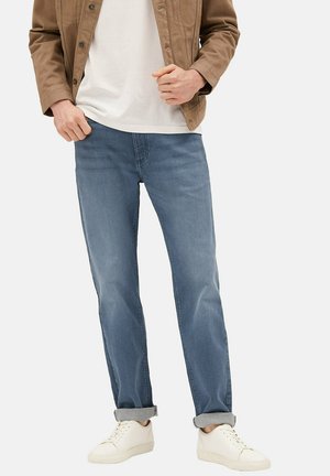 Muž stojící před jednobarevným pozadím, má na sobě vyhrnuté modré džíny, bílé tenisky, bílé tričko a hnědou bundu.