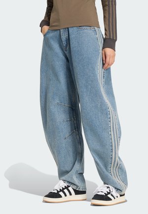 Person trägt lockere hellblaue Jeans mit Seitennähten und Streifen, kombiniert mit schwarz-weißen Sneakers und einem braunen Langarmshirt mit Streifendetails.