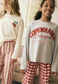 Zwei Mädchen tragen Freizeitkleidung: eines trägt ein cremfarbenes Oberteil mit Kirschstickerei und gestreifte Hosen; das andere trägt einen grauen Sweatshirt und rot karierte Hosen.