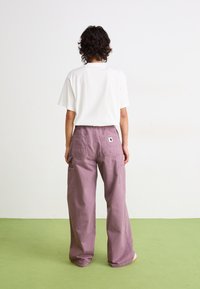 Biały t-shirt z krótkim rękawem w połączeniu z luźnymi, jasnofioletowymi spodniami typu cargo z naszywanymi kieszeniami i metką z tyłu. Płaskie buty, zielona podłoga.