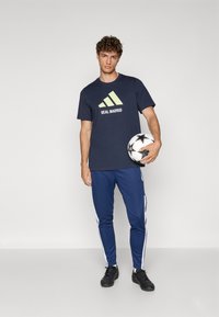 Mørkeblå trænings T-shirt med limegrøn Adidas-logo og "REAL MADRID" tekst; parret med blå træningsbukser med hvide sidestriber.