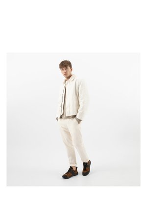 Młody mężczyzna stojący z rękami w kieszeniach, ubrany w jasnobeżową kurtkę, pasujące spodnie, szary sweter i brązowo-czarne trampki na białym tle.