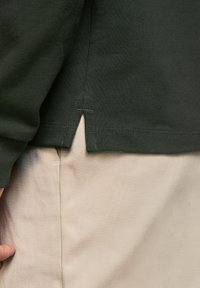 Camisa de manga larga verde oscuro con tejido texturizado, detalle de dobladillo dividido. Combinada con pantalones beige que tienen un ligero brillo y un patrón diagonal.
