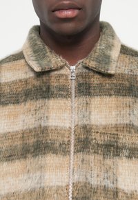 Plaid zip-up jacket in light brown, tan, and green. Made from textured fabric with a soft finish and a shirt collar. Silver zipper detail.
