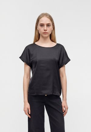 Jeune femme aux cheveux blonds raides portant un haut noir ample à manches courtes et un pantalon noir, debout devant un fond clair uni.