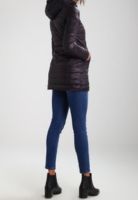 Chaqueta negra acolchada con capucha, que presenta mangas largas, bolsillos con cremallera y una silueta ligeramente más larga. Se lleva con jeans ajustados de color azul oscuro y botas negras de tobillo.