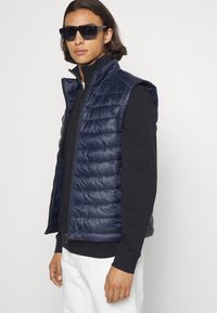 Vestă puffer marină cu textură matlasată, guler înalt și închidere cu fermoar, purtată deasupra unei cămăși negre cu mânecă lungă, completată cu ochelari de soare închiși la culoare.