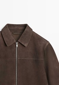 Giacca in suede marrone scuro con colletto a punta e zip frontale argentata, mostrata distesa su uno sfondo bianco.