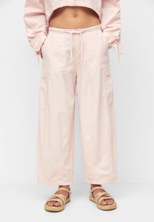 Personne portant un pantalon large rose clair avec taille à cordon et des sandales style corde, mains dans les poches, veste courte visible.