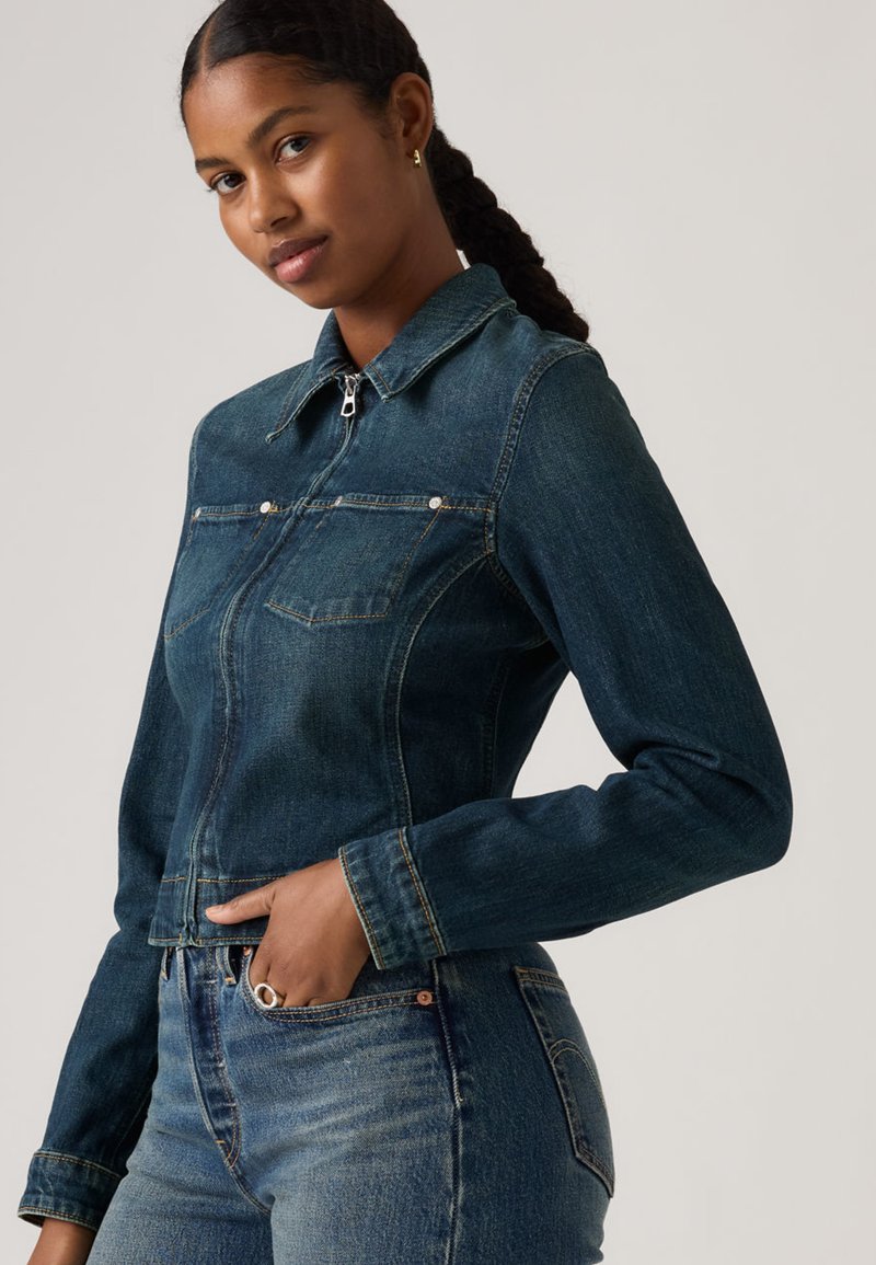 Γυναίκα με πλεξούδες, που φοράει σκούρο μπλε τζιν μπουφάν και τζιν παντελόνι, με το χέρι στην τσέπη, στέκεται μπροστά σε απλό φωτεινό φόντο.