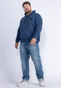 Felpa hoodie blu navy con cappuccio regolabile e logo frontale, abbinata a jeans azzurri e scarpe da ginnastica bianche. L'outfit offre una vestibilità comoda e uno stile informale.