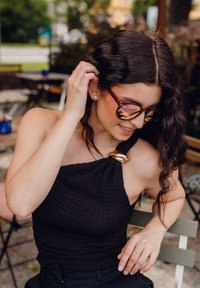 Černý texturovaný top se zlatým akcentem, oversized sluneční brýle s reflexními čočkami, vlnité tmavé vlasy a stříbrný prsten na ruce.