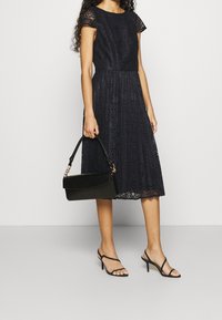 Robe en dentelle marine à manches courtes, corsage ajusté et jupe évasée. Associée à un sac à main noir et des sandales à talons fines noires.