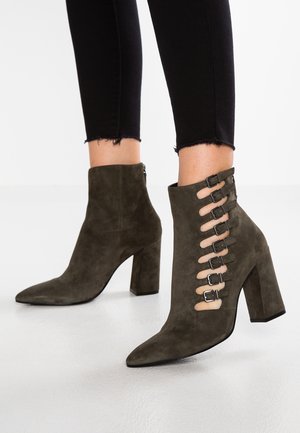 Bottines en daim vert olive à talons larges avec découpes latérales, fermées par sept petites boucles, portées avec un pantalon noir court.