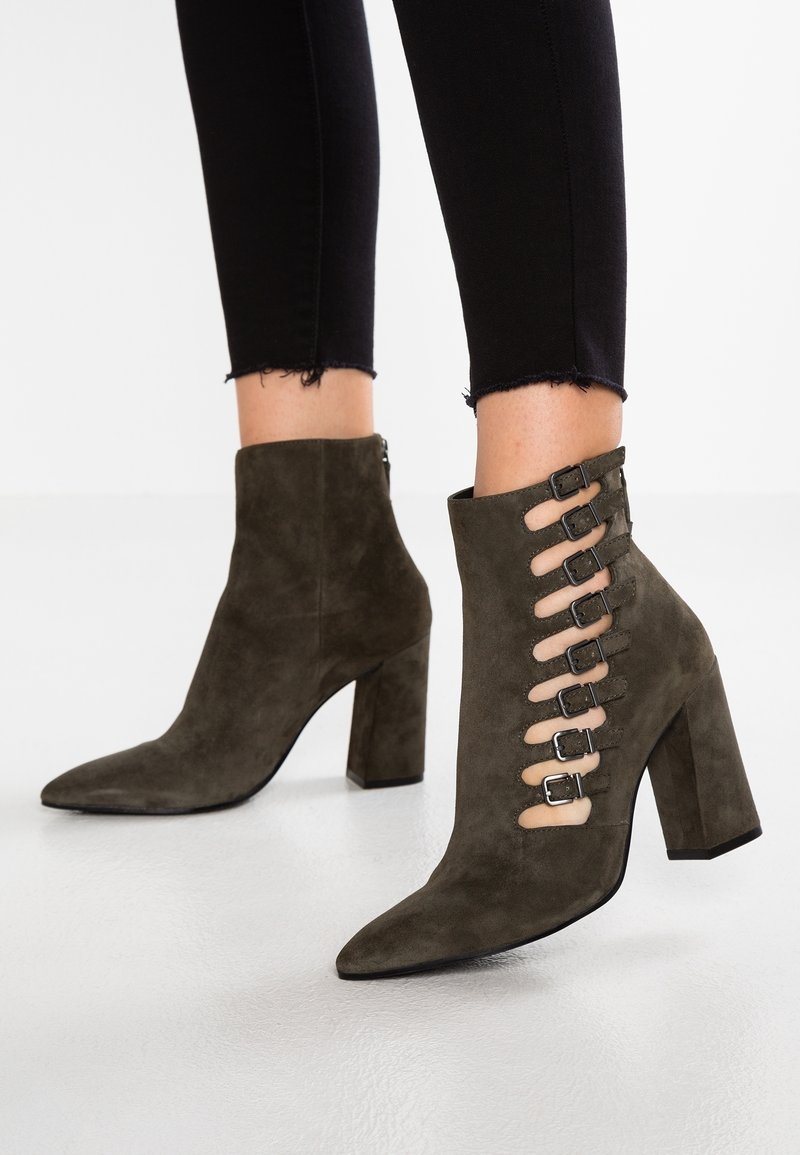 Bottines en daim vert olive à talons larges avec découpes latérales, fermées par sept petites boucles, portées avec un pantalon noir court.