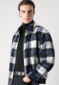 Fleece jacket with a black, white, and gray plaid pattern, accented with blue lines. Features a zip closure and side pockets.