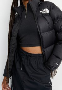 Svart puffjacka med dragkedja fram, hög krage och "The North Face"-logotyp på vänster bröst. Bärs över en åtsittande svart topp och byxor.