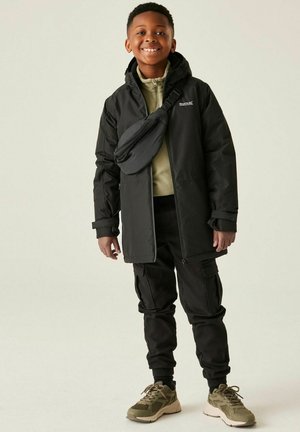 Uśmiechnięty chłopiec w czarnej kurtce parka, oliwkowo zielonym swetrze na zamek, czarnych spodniach typu cargo, torbie na ramię oraz beżowych sportowych butach stoi na tle jednolitego tła.