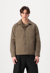 Brown, long-sleeve jacket with a shirt collar, front button closure, and two chest pockets. Worn over a matching top and black shorts.