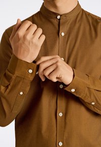 Camisa marrón de botones con cuello alzado, que presenta botones blancos, costuras visibles y una tela texturizada. Diseño de corte slim.